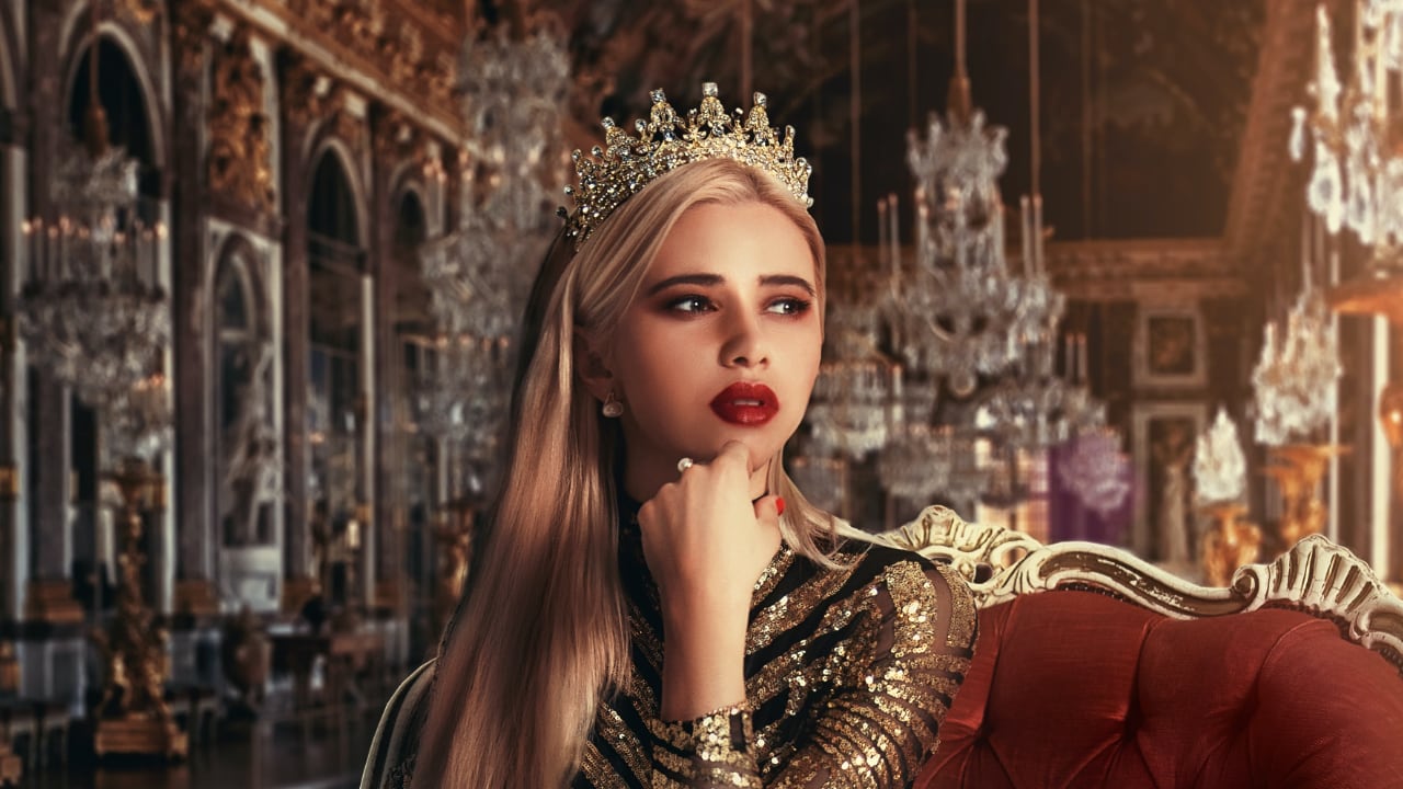 A blonde-haired woman with bright red lipstick wearing a crown sits on a couch in a royal room.